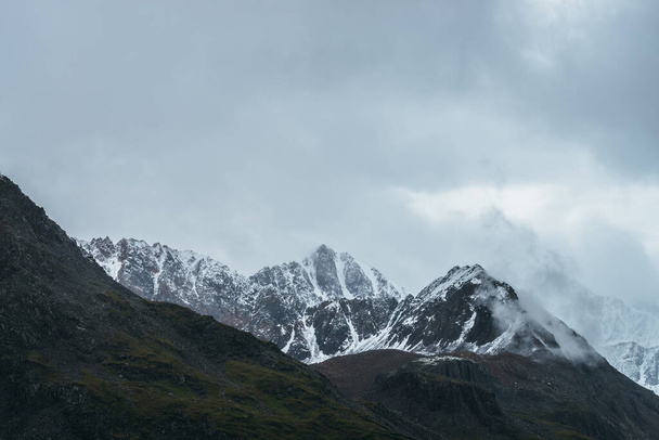 Paysage alpin atmosphérique avec une grande montagne culminant avec de la neige dans les nuages bas. Paysage montagneux dramatique avec apogée forte par temps couvert. Vue imprenable sur un pic pointu enneigé dans un ciel nuageux. - Photo, image