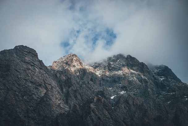 Atmospheric mountain landscape with low cloud on mountain top in sunlight. Dark rocks with snow and blue skylight in gray cloudy sky. Beautiful mountain scenery with low cloud on rocky top in sunshine - Photo, image