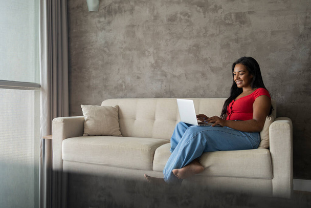 Young black woman working at home with laptop on her lap sitting on her couch in the living room.  Home office concept. Gray notebook for working. High quality photo - Zdjęcie, obraz