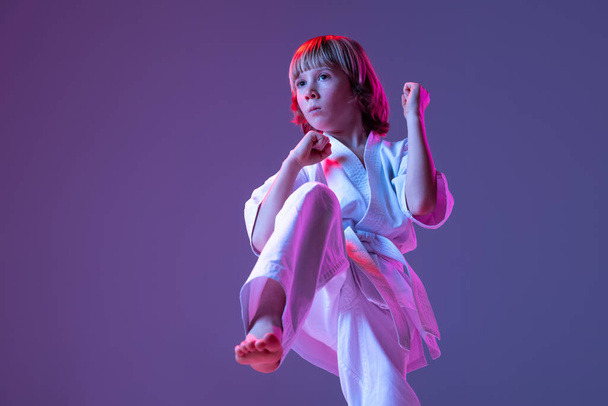 Dynamisch portret van kind, jonge mannelijke taekwondo, karate atleten in doboks doen basisbewegingen geïsoleerd op paarse achtergrond in neon. Begrip sport, vechtsport - Foto, afbeelding