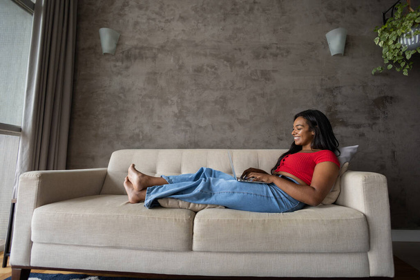 Egy fiatal fekete nő, aki otthon dolgozik, laptoppal az ölében fekszik a kanapéján a nappaliban. Otthoni iroda koncepció. Szürke jegyzetfüzet a munkához. Kiváló minőségű fénykép - Fotó, kép