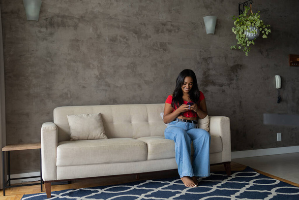 Evinde akıllı telefonuyla çalışan genç siyahi bir kadın oturma odasında kanepesinde oturuyor. Merkez ofis konsepti. Yüksek kalite fotoğraf - Fotoğraf, Görsel