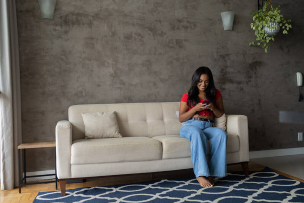Joven mujer negra trabajando en casa con smartphone sentado en su sofá en la sala de estar. Concepto de oficina en casa. Foto de alta calidad - Foto, Imagen