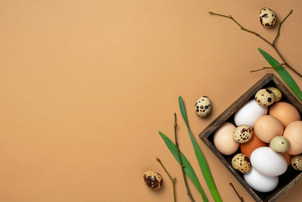 Easter flat lay in trendy color. Chicken and quail eggs in wooden box with branches and green leaves on beige background. Top view, copy space - Fotó, kép