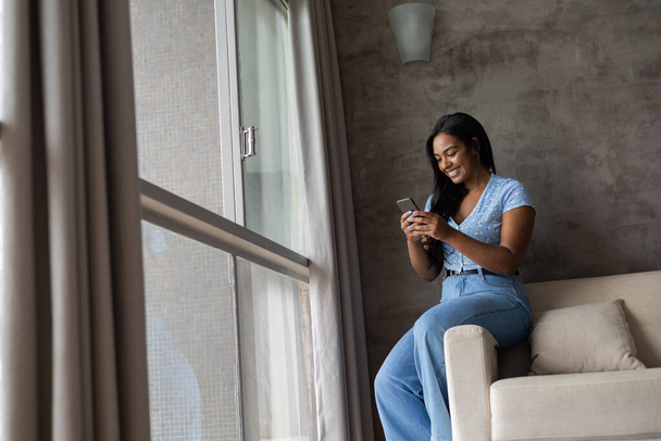 Young black woman working at home with smartphone sitting on her couch in the living room.  Home office concept. High quality photo - Photo, image