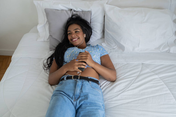 Young black woman in her bed listening to music with headphones at home inside her apartment. Feel good concept, happy. High quality photo.  - Foto, Bild