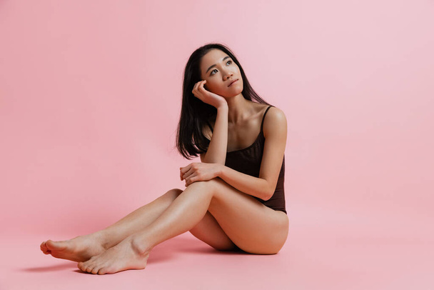 Studio portrait of dreamy looking tender woman sitting and posing in bodysuit isolated over pink background - Foto, immagini