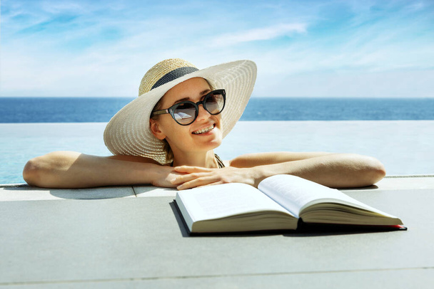 smiling attractive woman with hat and sunglasses reading a book and relaxing in infinity swimming pool at vacation resort - Photo, Image