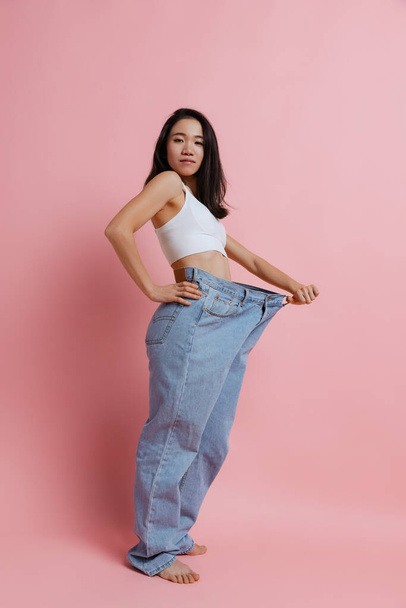 Full-length portrait of beautiful slim woman posing in huge jeans isolated over pink studio background. Weight loss concept - Foto, immagini