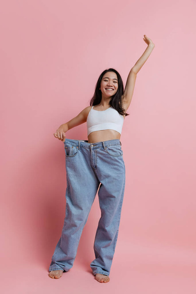 Full-length portrait of beautiful slim woman posing in huge jeans isolated over pink studio background. Weight loss concept - 写真・画像