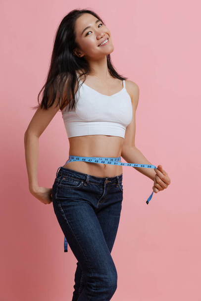 Portrait of slim woman measuring waist with tape, posing isolated over pink studio background. Weight loss concept - 写真・画像