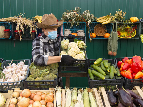 Chou-fleur au marché fermier Un vendeur masculin place une boîte de chou-fleur sur le comptoir Le vendeur dans un masque médical protecteur. - Photo, image