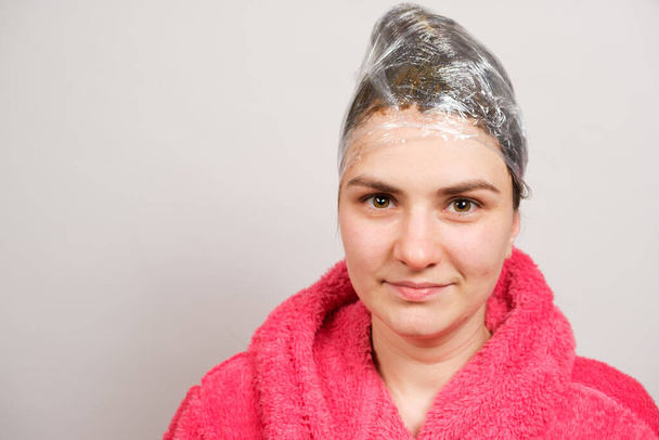 Een mooie brunette wikkelt haar hoofd met een transparante film, een wrap en een haarmasker, een spandoek met een plek voor tekst - Foto, afbeelding
