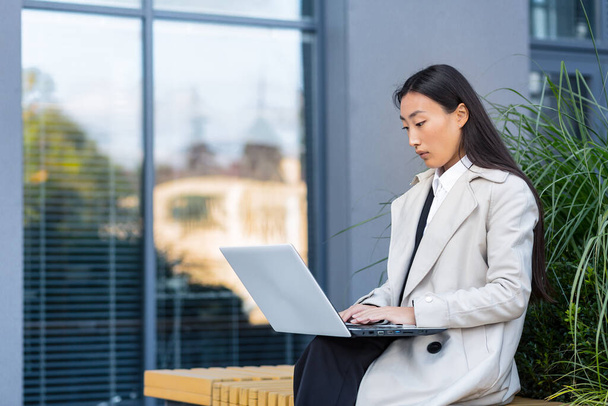 Asian business woman working with laptop outside office, sitting on bench during work break - 写真・画像