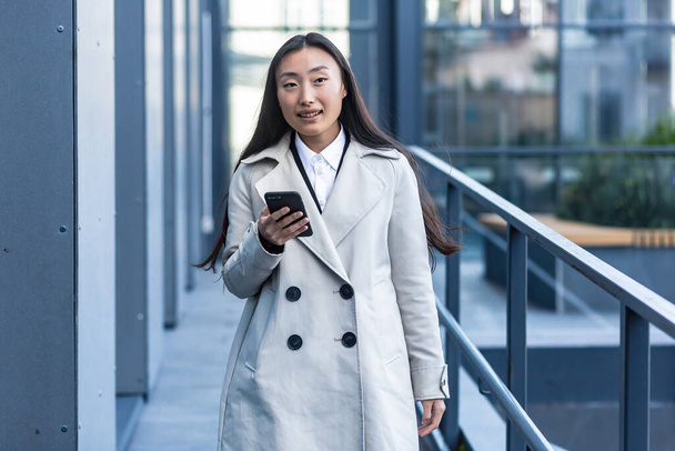 Gyönyörű ázsiai nő üzleti ruhában használja a telefont, sétál közel a modern irodaház kívül - Fotó, kép