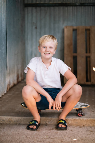 Cute blonde pre-teen boy with relaxed pose sitting in rustic outdoor county setting - Photo, Image