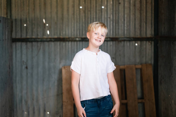 Cute blonde pre-teen boy with relaxed pose sitting in rustic outdoor county setting - Foto, afbeelding