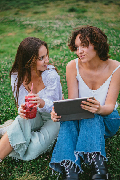 Two friends looking at each other while they are checking on social media on a tablet computer. They are sit on the grass. - Photo, Image