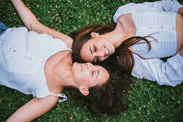 Two teenager girls smiling and looking at each other while they are laying down on grass in a park. They seem relaxed. - Photo, image