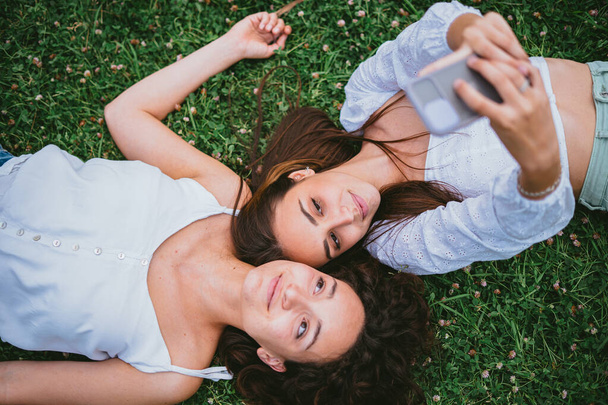 Two friends taking a selfie photo while they are laying down on the grass in a park. - Foto, imagen