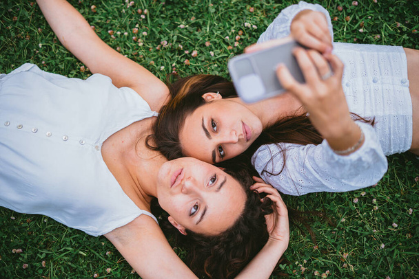 Two teenager girls posting a photo on social media. They are laying down on the grass in a park. - Photo, image