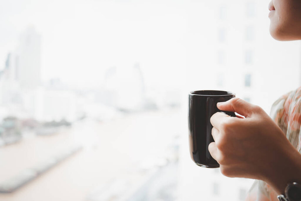 Young Asian woman holding a coffee cup near the window, woman drinking a coffee. Happy woman drinking hot coffee in a mug in the morning close up with copyspace. - Fotó, kép