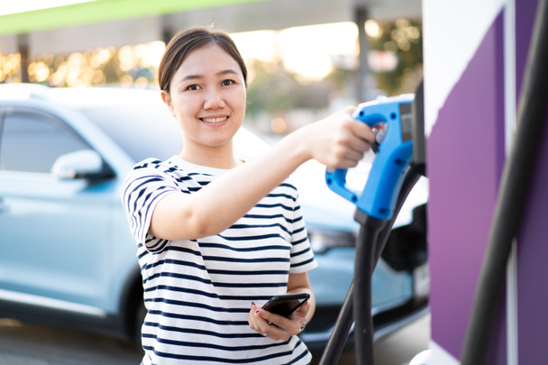 Happy Asian woman holding a DC - CCS type 2 EV charging connector at EV charging station, woman preparing an EV - electric vehicle charging connector for recharge a vehicle. - Foto, afbeelding