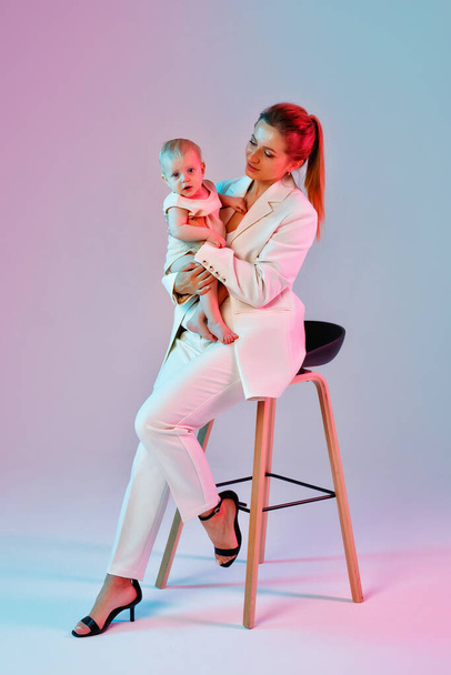 Una joven con un traje ligero está sentada en una silla sosteniendo a un bebé en sus brazos. Una mujer de negocios con un bebé en una mezcla de neón rosa y azul claro. - Foto, Imagen