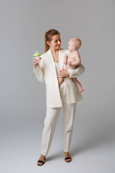 Kevytpukuinen liikenainen pitää vauvaa ja maitopulloa sylissään. Äiti ja vauva studiossa harmaalla taustalla. - Valokuva, kuva