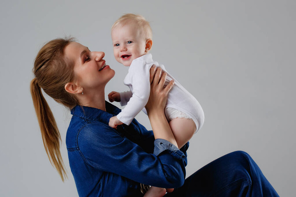 A woman in a denim jumpsuit holds a child in a white bodysuit. Mother and son in the studio on a gray background. Motherhood concept. Unconditional love. - Фото, зображення