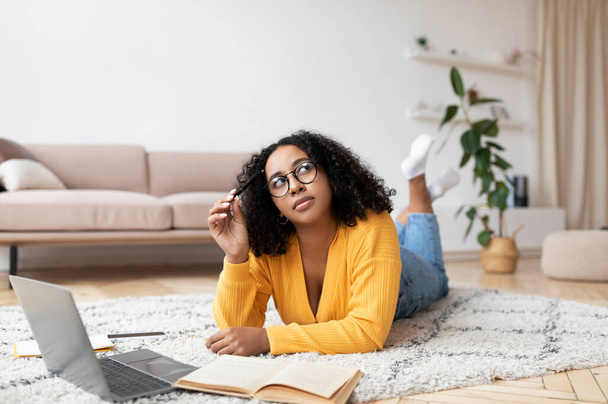 Távoktatási koncepció. Átgondolt afro-amerikai hölgy használja laptop és olvasókönyv, feküdt a padlón otthon - Fotó, kép