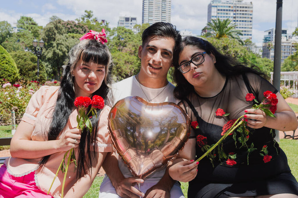 Két fiatal latin latin latin kaukázusi lány és egy fiú, egy nyilvános parkban ülnek együtt, virágokkal a kezükben, és egy szívlufi nézi a kamerát. a szeretet és a barátság fogalma. - Fotó, kép