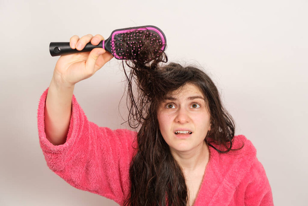 Μια γυναίκα προσπαθεί να χτενίσει τα μπερδεμένα βρεγμένα μαλλιά της. Φροντίδα μαλλιών στο σπίτι, τριχόπτωση και τμήμα. - Φωτογραφία, εικόνα