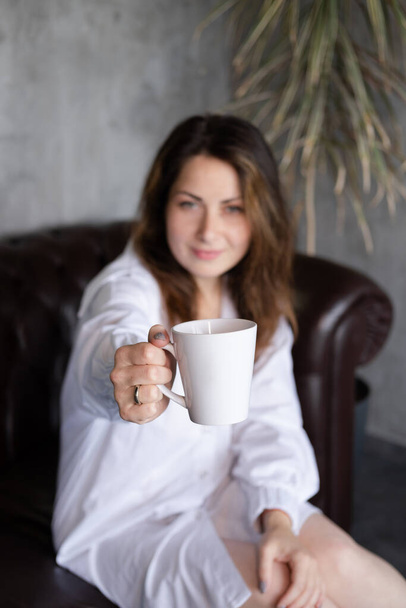 woman in white shirt on leather couch holding white cup. focus on mug. mock up. copy space - Foto, Imagem