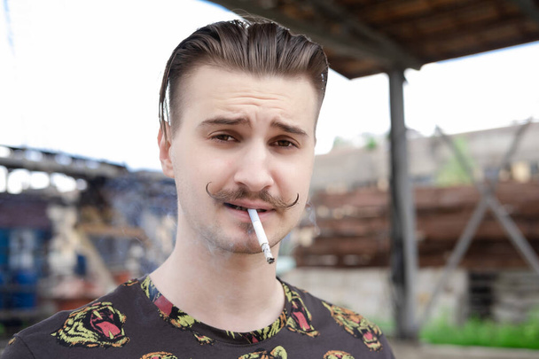 Nuori, itsevarma mies, jolla on viikset ja joka polttaa tupakkaa ulkona. huono tapa, tupakkariippuvuus - Valokuva, kuva