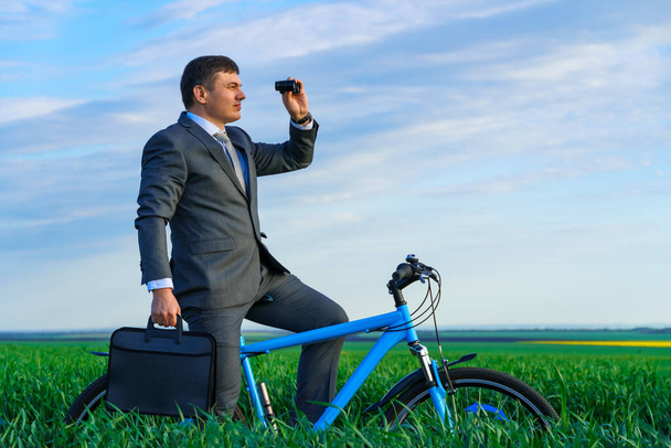 concept d'entreprise - Un homme d'affaires fait du vélo sur un champ d'herbe verte, regarde au loin à travers des jumelles, vêtu d'un costume d'affaires, il a une mallette et des documents - Photo, image