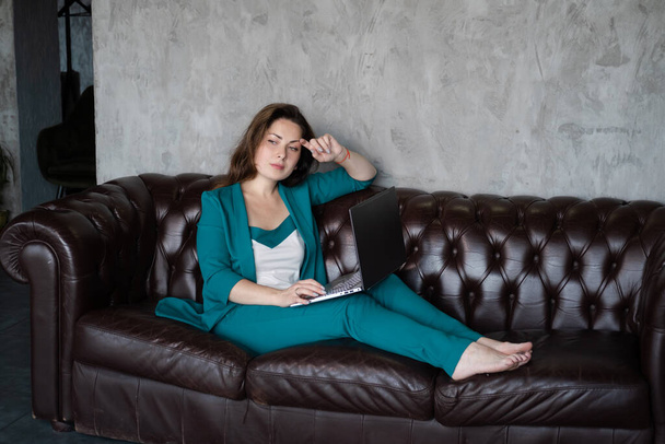 遠くの仕事、遠隔研究。自信のあるブルネットの女性ラップトップ付きの革のソファの上に裸足で緑のスーツ。内務省だ。家から仕事で - 写真・画像