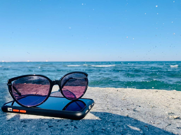 Sunglasses laying on a phone on stone near the sea - Foto, afbeelding