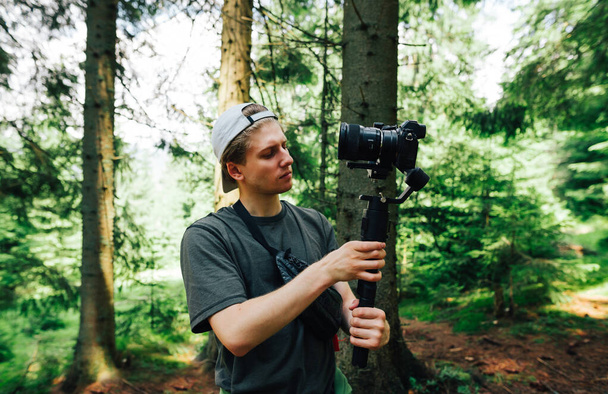 Un joven camarógrafo con una cara seria se encuentra en un bosque de montaña con una cámara en el estabilizador y ajusta el equipo para disparar. Crea contenido de viajes para tu blog. - Foto, Imagen