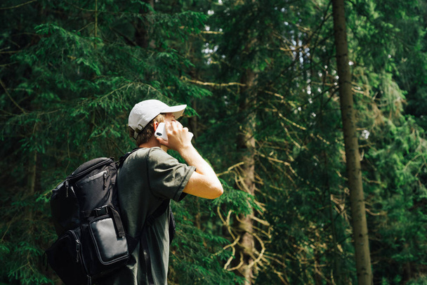 Genç hippi bir adam dağ yürüyüşünde telefonla konuşuyor, ormanda duruyor, akıllı bir telefonla iletişim kuruyor, başını çeviriyor, sırtında bir sırt çantası ve bir şapka taşıyor. - Fotoğraf, Görsel