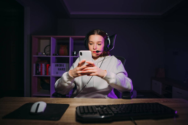 Attractive female streamer sitting at night at the computer in a headset, playing games, using a smartphone during a break in a room with purple light. - Фото, изображение