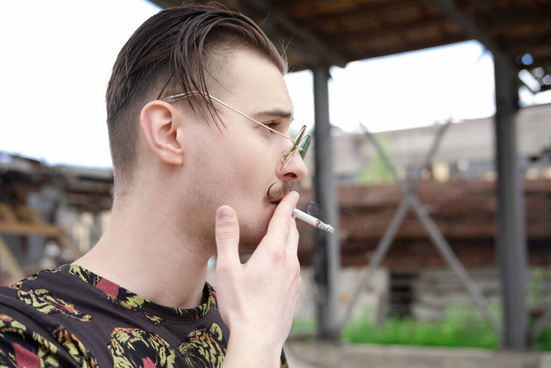 joven hombre confiado con bigote fumando un cigarrillo afuera. mal hábito, adicción al tabaco - Foto, imagen