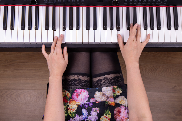 Un joli modèle féminin mince portant des bas et de la lingerie sous une robe fleurie tout en s'asseyant et en jouant un piano avec ses doigts sur les touches du piano - Photo, image