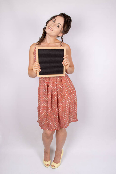Привлекательная модель с длинными коричневыми рыжими волосами на белом фоне в винтажном платье с пустой вывеской - Фото, изображение
