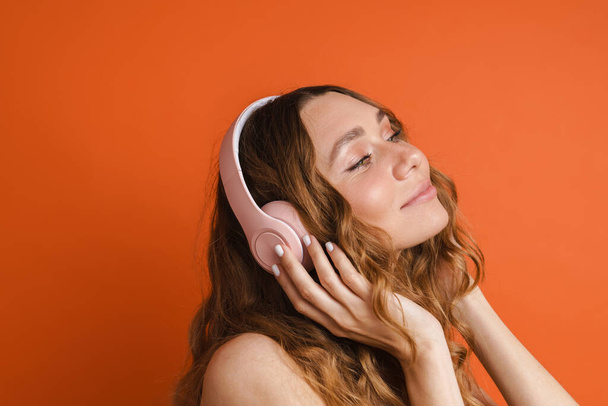 Young ginger woman smiling while listening music with headphones isolated over orange wall - Fotografie, Obrázek