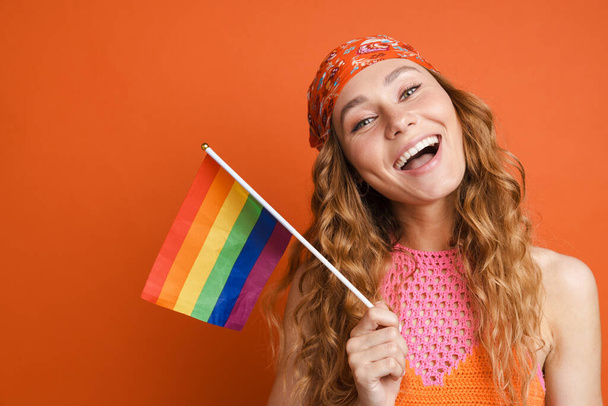 Young ginger woman in bandana smiling while posing with rainbow flag isolated over orange wall - Fotoğraf, Görsel