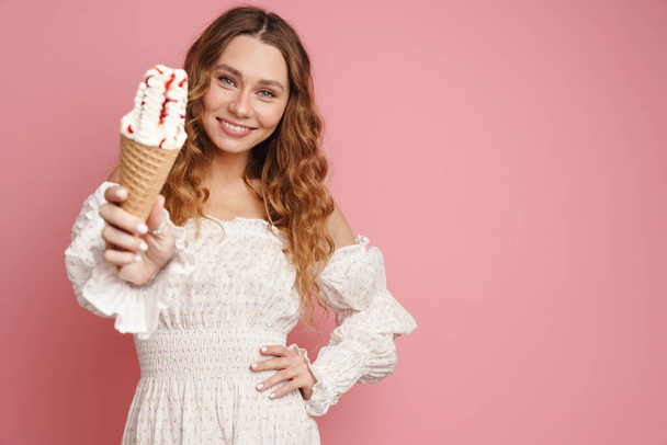 Giovane donna zenzero in abito sorridente mentre mostra il gelato isolato su parete rosa - Foto, immagini