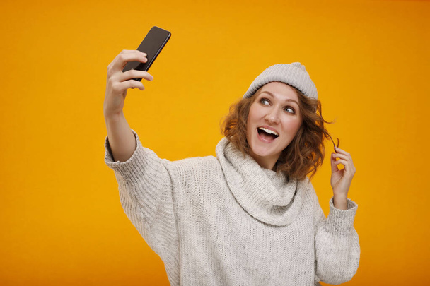 Young happy woman with curly hair in winter hat standing over yellow background making selfie. Happy New Year and Christmas concept. Place for your text. - 写真・画像