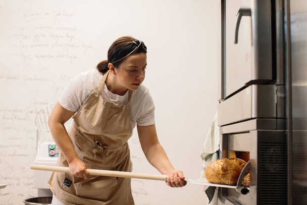 Female baker using a peel to take out a loaf of bread of the oven in a bakery - 写真・画像