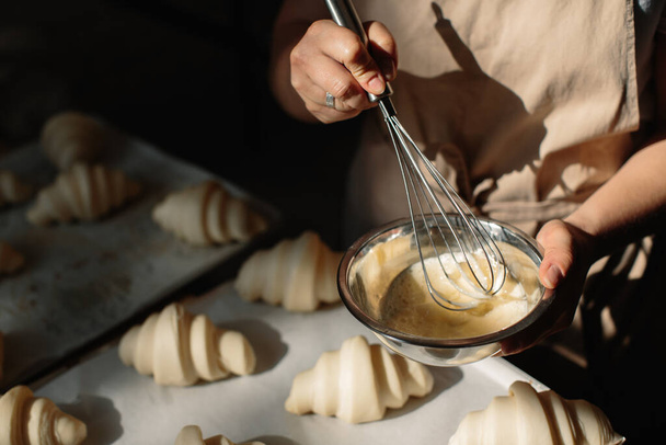 Las hembras del panadero untan cruasanes crudos con un cepillo en la yema, el proceso de cocción. - Foto, Imagen
